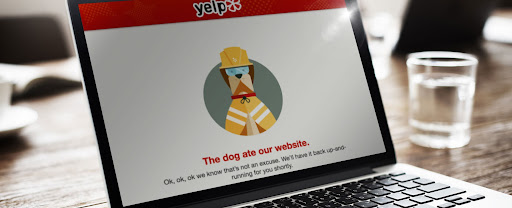 A laptop on a table opened up to a Yelp empty state page with an illustration of a dog wearing a hardhat with the text “The dog ate our website” below.