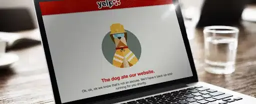 A laptop on a table opened up to a Yelp empty state page with an illustration of a dog wearing a hardhat with the text “The dog ate our website” below.